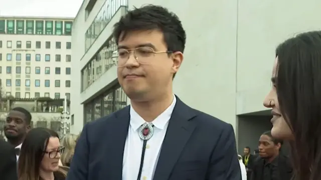 A man in a suit jacket, white shirt and bolo tie with a red jewel in the centre stands in a crowd, looking on as an interviewer asks a question.