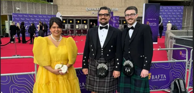 A woman in a yellow dress with large exaggerated shoulders stands next to two men in Scottish kilts, both wearing smart jackets and bow ties. Behind them is a red carpet and signage displaying the Bafta Game Awards gold mask