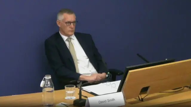 David Smith pictured sitting at a desk wearing glasses while being questioned at the inquiry.