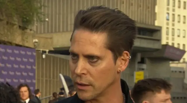 A man with slicked back hair, eyeshadow and several ear piercings looks intensely at a reporter on the red carpet