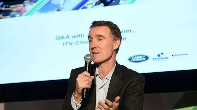 Adam Crozier speaks at a conference while holding a microphone and a World Rugby screen appears behind him.