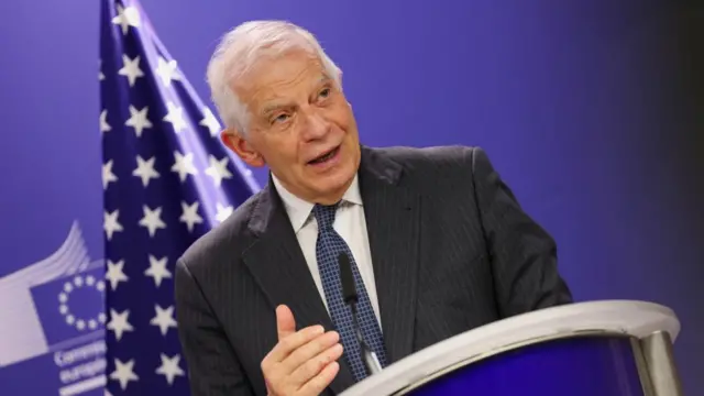 Josep Borrell stands at a podium in a blue tie and black suit