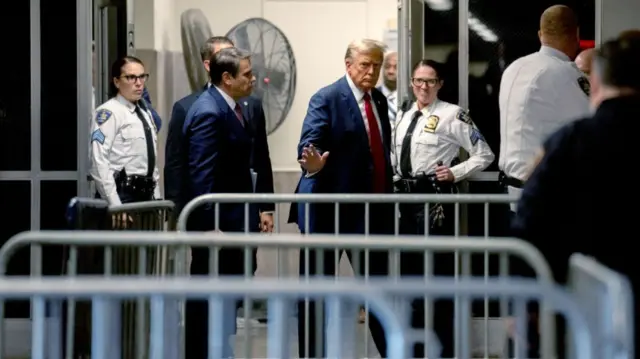 Donald Trump is seen walking outside of a courtroom, waving with his right hand, surrounded by security officers. He wears a red tie and blue suit