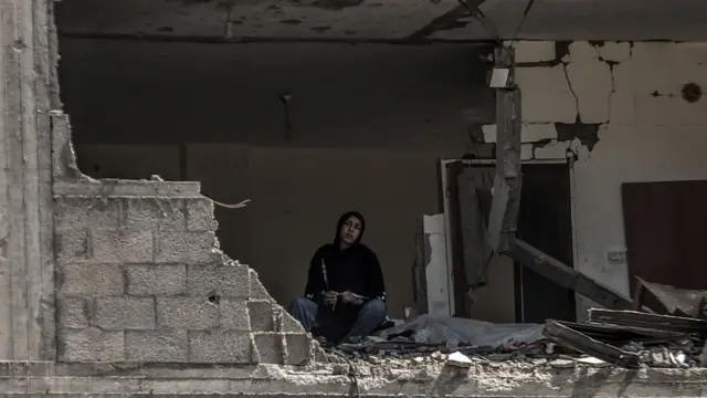 A woman sits in an apartment, the exterior wall of which has been blown up, leaving the inside exposed