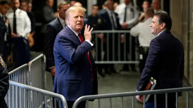 Donald Trump gestures with his right hand while standing outside a New York courtroom while he is boxed in by metal fences and he stands beside another man in a dark suit, who smiles at him.