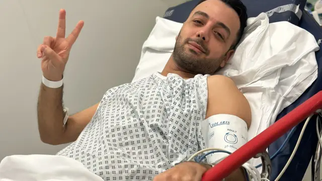 Iranian TV presenter Pouria Zeraati lies in a hospital bed, wearing a gown. He makes a peace sign with one hand