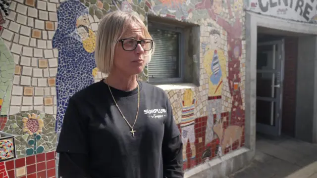 Claire Hopkins, a charity co-founder, speaks to a camera and is dressed in a black t-shirt and stands in front of a colourful building