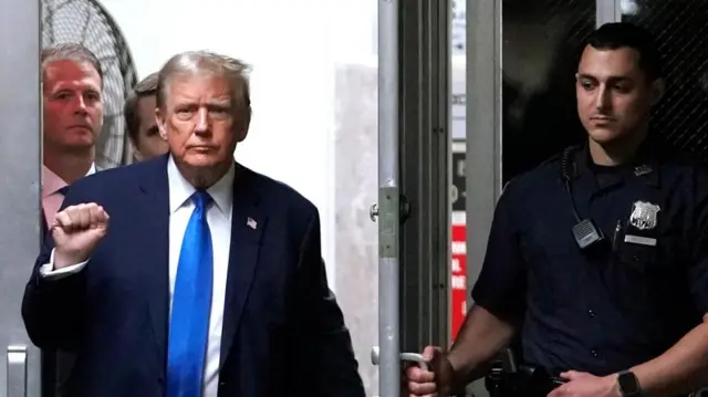 Donald Trump re-enters the courtroom and raises a fist, dressed in a blue suit and a blue tie.