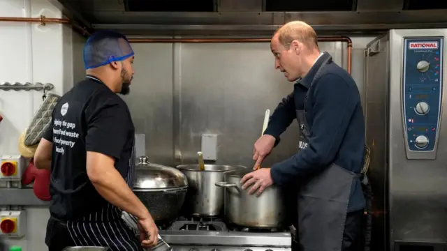 Prince William stirring food in a pan while a chef talks to him