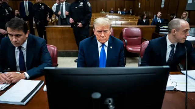 Donald Trump sits at a brown table in a courtroom while his attorneys sit on either side of him. He wears a blue suit and blue tie and appears to scowl at the camera