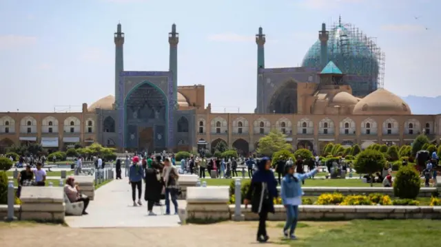 People walking in Isfahan's Naqsh-e Jahan Square, with the turquoise dome and minaret's of the city's 17th century Shah Mosque in the background