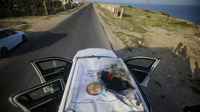 Image of the roof of a car with a large hole in it, part of a logo saying "World Central Kitchen" is clearly visible and partially destroyed.