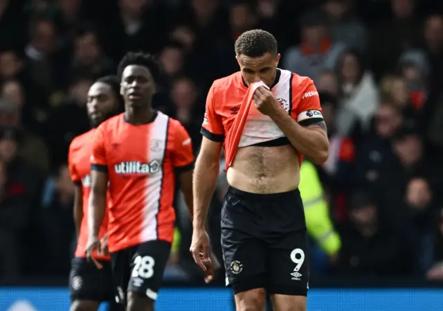 Carlton Morris covers his face with his shirt as Luton trudge back to kick off