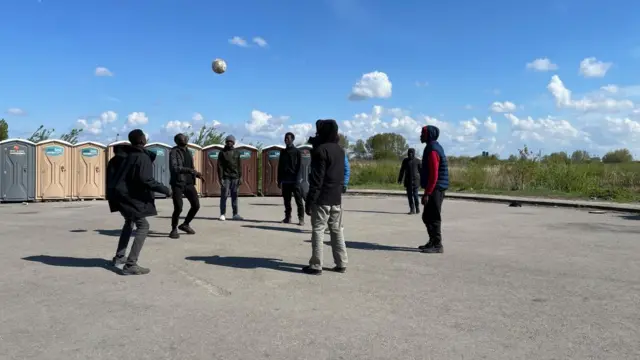 Migrants in Calais play football in a parking lot beside portapotties.