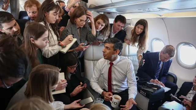 Rishi Sunak with a coffee cup in his hand in an aircraft surrounded by a group of reporters