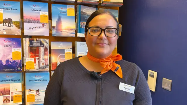 A woman in travel agent uniform stands in front of a wall of brochures