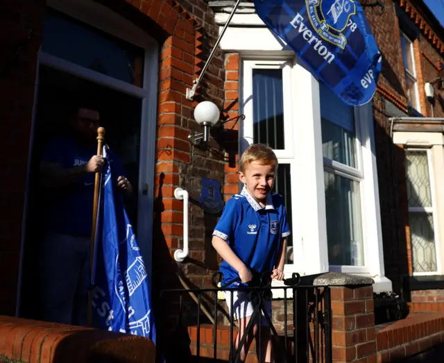 A young Eveerton fan sits atop the gate to his house as his dad carries a flag out of the front door