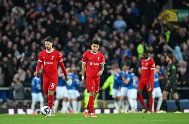 Liverpool players trudge back to kick off as Everton celebrate with their fans in the background