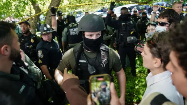 A police officer in a mask and helmet approaches a group of people, one of whom films him