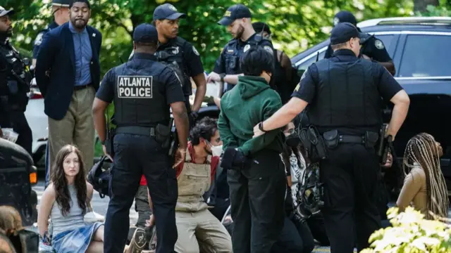 Five people sit on the ground, handcuffed, while police arrest others around them