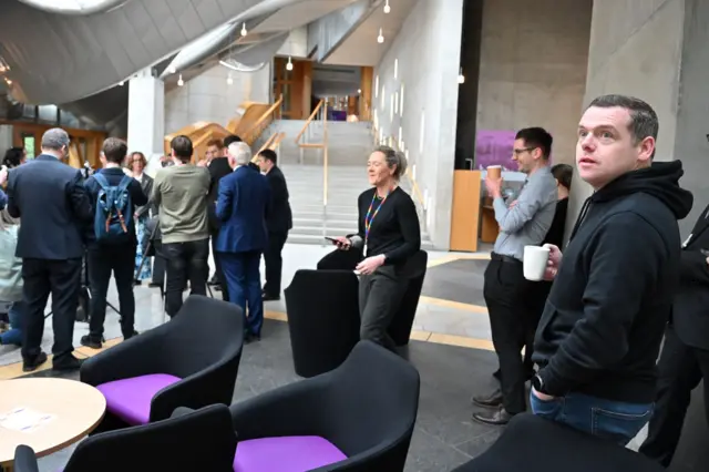 Crowd of journalists and Douglas Ross watching from the side holding a coffee cup