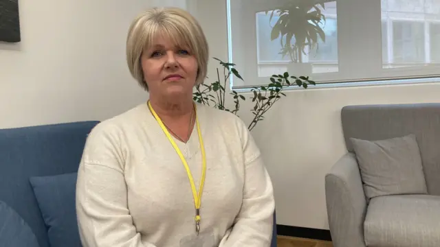 Nicki, a woman with short blonde hair, looks at the camera as she sits on a sofa