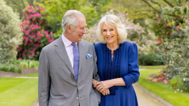 King Charles, in a grey suit and blue dotted tie, and Queen Camilla, who is wearing a blue dress, link arms while standing in the palace gardens.