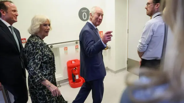 King Charles points and smiles at hospital staff while visiting a cancer centre in central London while Queen Camilla stands beside him on the left