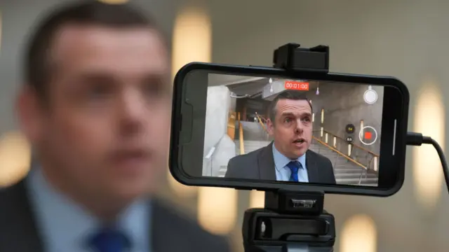 In the foreground is phone strapped to a black tripod with the screen showing Douglas Ross being recorded, and the background shows a blurry Douglas Ross talking to the camera