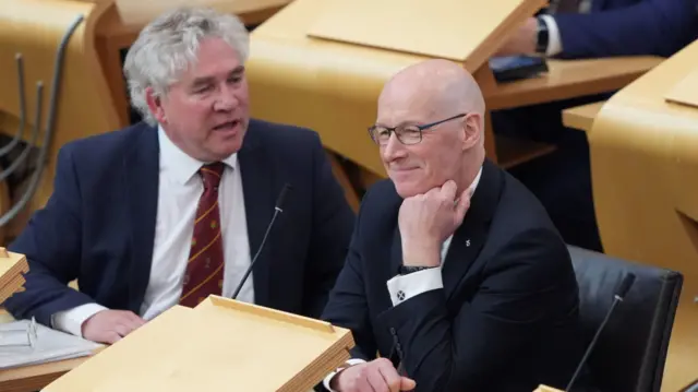 SNP MSP Kenneth Gibson talking to a smiling Former Deputy First Minister John Swinney as they are both seated in parliament.