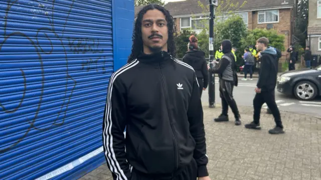 Diogo, a young man with long hair, a moustache and wearing a black jacket, looks at the camera