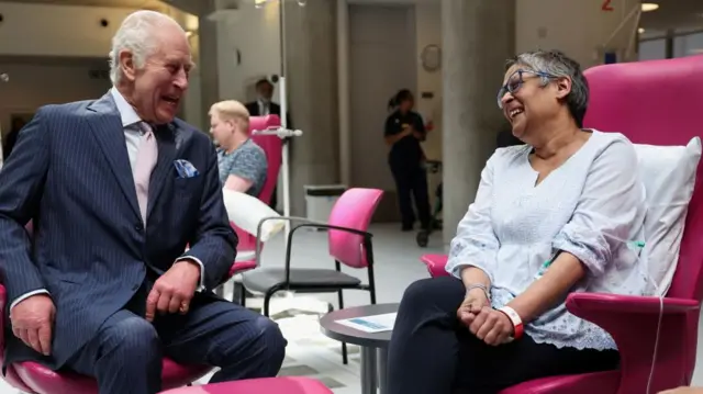 Charles and a woman with a drip in her arm sit in chairs, smiling and laughing at each other