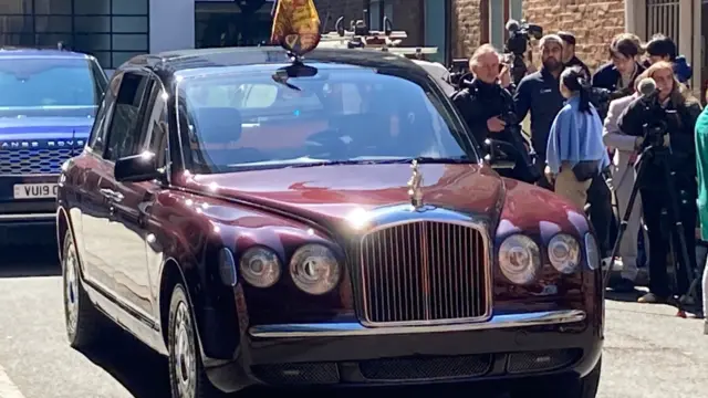 A deep red Bentley, flying the royal standard, on a road flanked by cameras and reporters