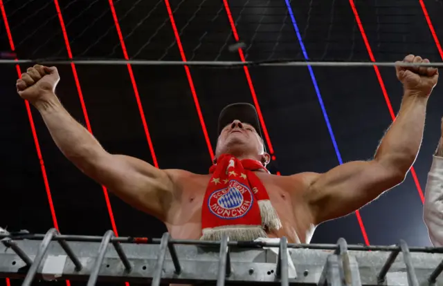 A shirtless Bayern fan wearing a club scarf gestures support from the stands