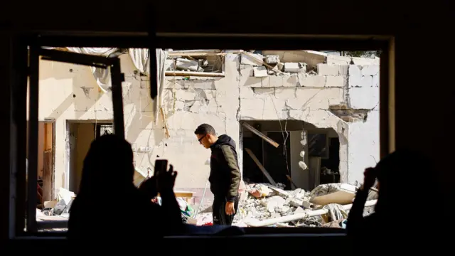 A woman in the shadows looks out a window at the rubble of a destroyed house in Rafah