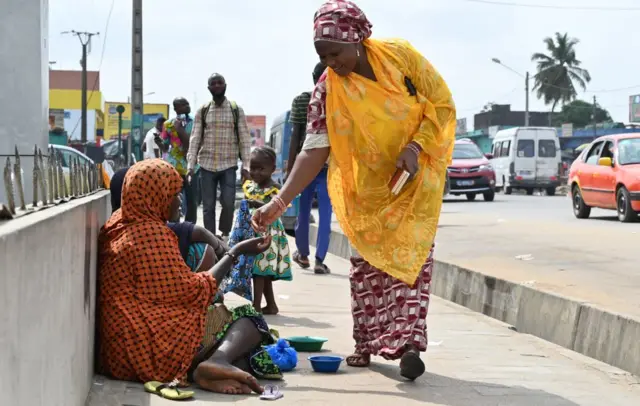 A pedestrian gives mony to a Nigerien woman sitting on the sidewalk with her children in Abobo suburb of Abidjan on March 28, 2022.