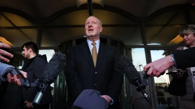 Alan Bates stands in front of microphones for a media appearance, dressed in a suit and tie