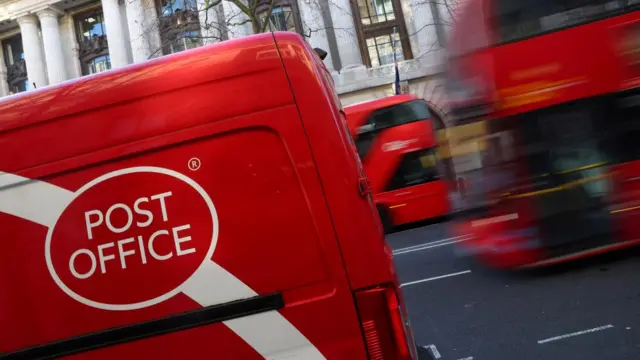 A red Post Office mailbox is seen in front of a blurred-out bus