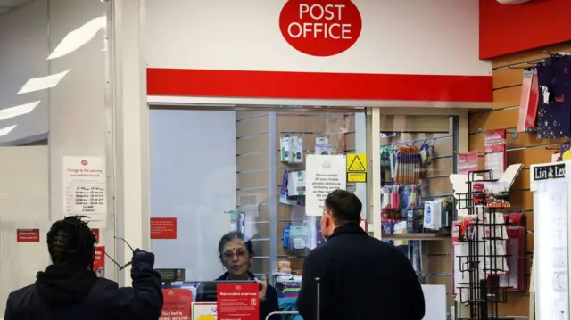 An image from inside a post office, where a sub-postmistress is seen behind a window-shielded kiosk serving a male customer. Another man stands to his far left in the queue.