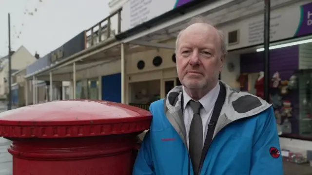 Alan Bates, dressed in a blue overcoat, stands beside a Post Office