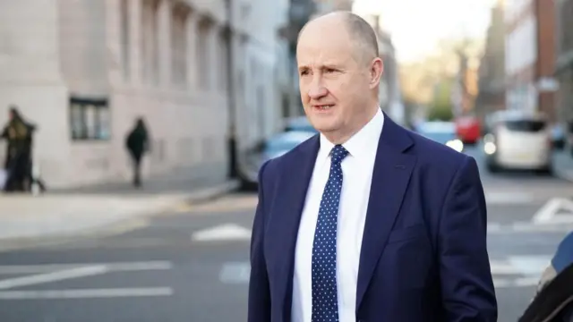 Post Office Minister Kevin Hollinrake walks across the street dressed in a blue suit and tie.