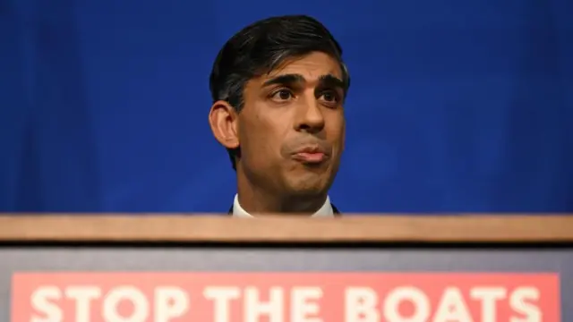 Rishi Sunak stands in front of a podium with the sign 'Stop the Boats' beneath it