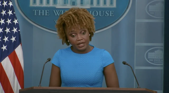 Karine Jean-Pierre, the White House Press Secretary, speaks at a podium in a blue dress with an American flag in the background