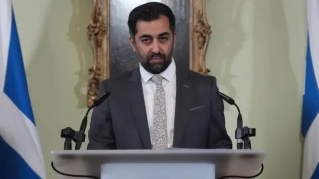 Yousaf speaks at a lectern, flanked by two Scottish flags.