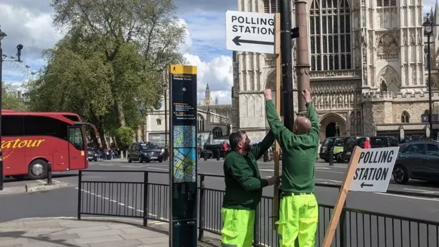 Construction workers affix a polling station sign to a pole and a red bus drives by in the background.