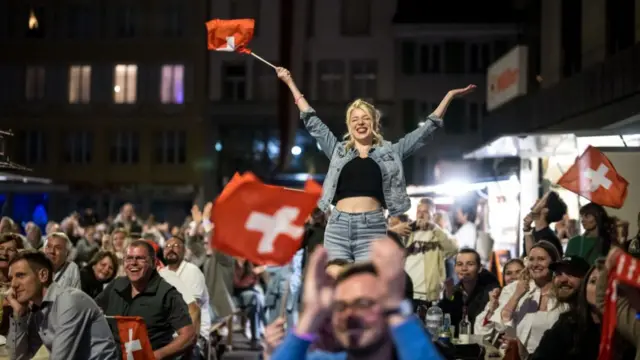 People holding the Swiss flag gather in the street in Biel, with a woman in a jean jacket standing on a table.