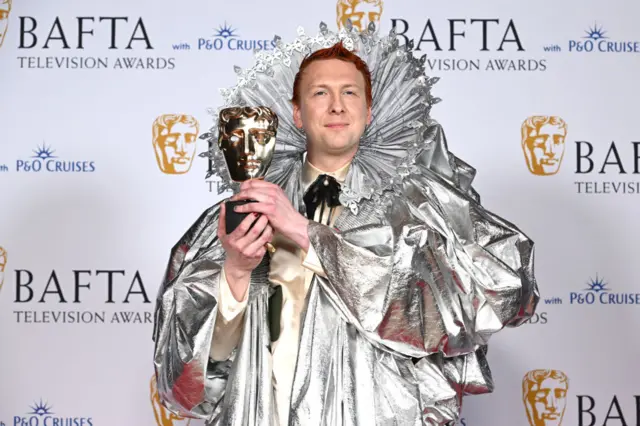 Joe Lycett dressed as Queen Elizabeth I holding his Bafta award