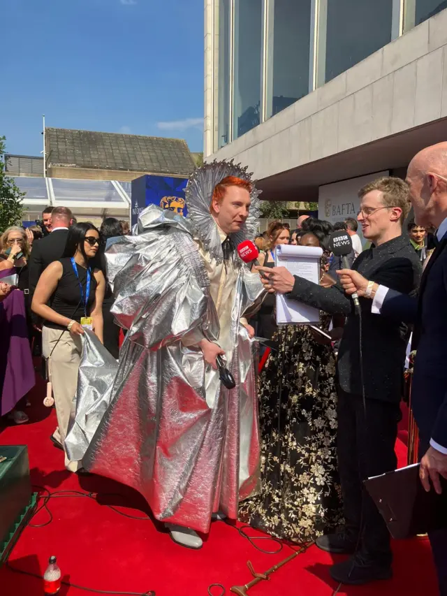 Joe Lycett dressed in a gown talking to BBC presenters