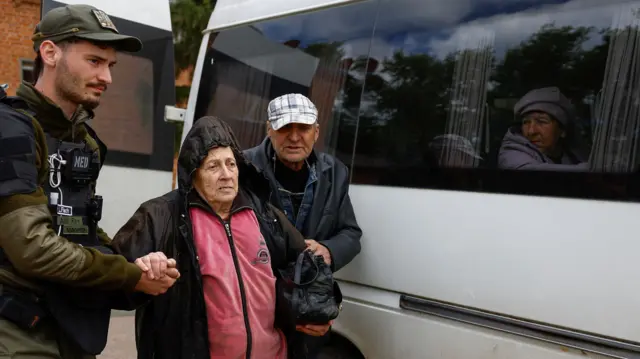 An volunteer helps an elderly man and woman to a bus where another elderly woman looks out of the window