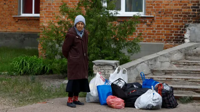 An elderly woman stands with numerous bags on the floor next to her, wearing a coat and blue hat